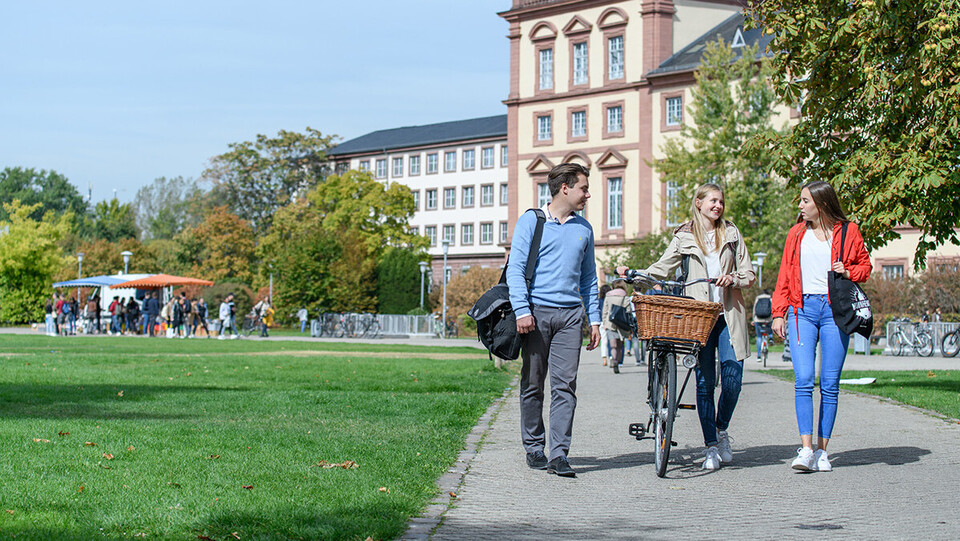 Drei junge Menschen unterhalten sich auf einem Gehweg vor dem Mannheimer Schloss im Mensapark, die Person in der Mitte schiebt ein Fahrrad.