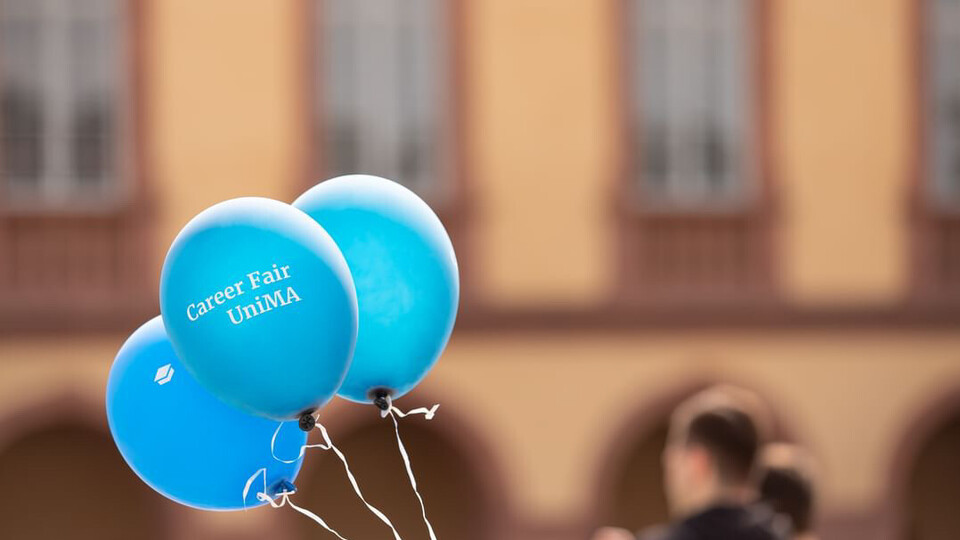 Drei blaue Luftballons, auf einem steht Carer Fair. Im Hintergrund sieht man verschwommen zwei Personen und das Schloss.