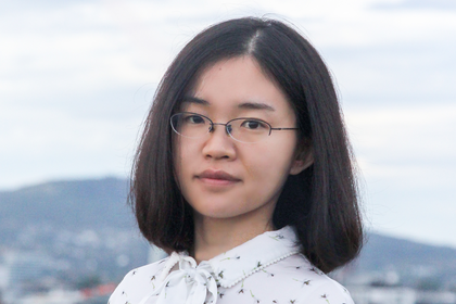 Lei Li im Freien, im Hintergrund sieht man einen Berg und eine Stadt. Sie trägt eine Brille und eine geblümte Bluse mit Schleife.
