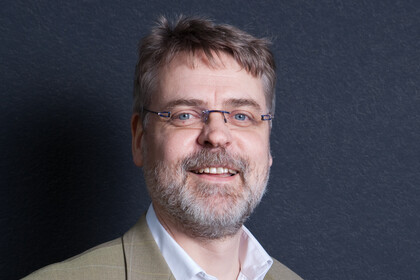 Portrait von Hartmut Wessler. Er steht in einem beigen Anzug vor einer dunkelgrauen Wand.