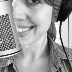 Black and white photo of a smiling woman in front of a microphone with headphones on her ears. 