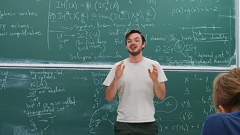Junger Mann steht vor einer Tafel und spricht zu einer Klasse