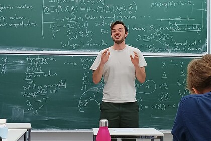 Junger Mann steht vor einer Tafel und spricht zu einer Klasse