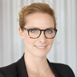 Portrait photo of a smiling blonde woman with a pinned-up hairstyle, horn-rimmed glasses, dressed in a black blazer and a gold chain around her neck.