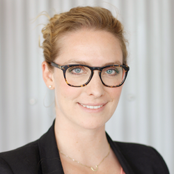 Portrait photo of a smiling blonde woman with a pinned-up hairstyle, horn-rimmed glasses, dressed in a black blazer and a gold chain around her neck. 