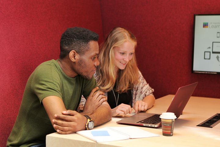 Two students are sitting next to each other in a red corner seat at a desk in the library. They are looking together at the computer in front of them. There is a coffee mug and an open book on the table next to the computer