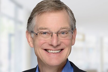 Die Person auf dem Porträtfoto trägt ein schwarzes Jackett, ein blaues Hemd und lächelt. Der Hintergrund ist unkenntlich gemacht.