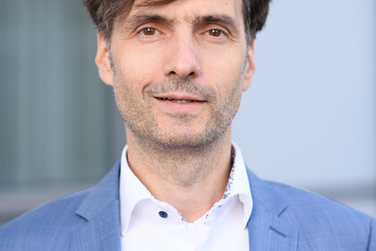 Portrait von Georg Alpers. Er trägt ein weißes Hemd sowie einen blauen Blazer und lächelt.