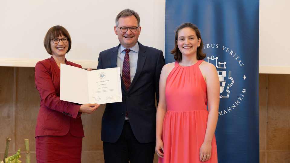 Prof. Gassert bekommt eine Urkunde überreicht. Im Hintergrund ein blaues Banner mit dem Logo der Universität Mannheim.