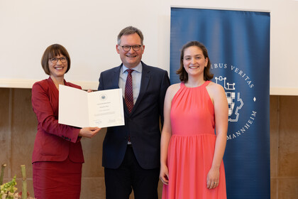 Prof. Gassert bekommt eine Urkunde überreicht. Im Hintergrund ein blaues Banner mit dem Logo der Universität Mannheim.