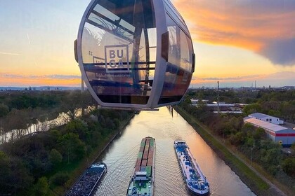 Eine BUGA-Gondel passiert den Fluss Neckar. Auf dem Fluss fahren gerade zwei große Lastschiffe. Im Hintergrund färbt sich der Abendhimmel rötlich. Link: Instagram Buga Mannheim 2023