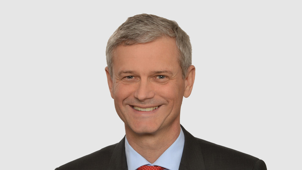 Prof. Helmig hat kurze graue Haare und trägt ein dunkles Jacket, ein hellblaues Hemd und eine rote Krawatte.