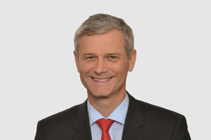 Prof. Helmig hat kurze graue Haare und trägt ein dunkles Jacket, ein hellblaues Hemd und eine rote Krawatte.