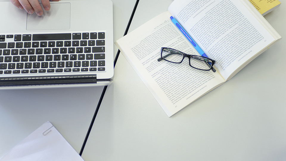Ein Laptop, ein aufgeschlagenes Buch und eine Brille liegen auf dem Tisch.