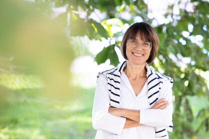 Prof. Dr. Annette Kehnel. Sie steht lächelnd unter einem Baum, dabei trägt sie eine Brille und eine weiße Bluse, ein gestreifter Pullover liegt um ihre Schultern.