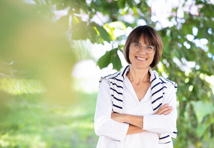 Prof. Dr. Annette Kehnel. Sie steht lächelnd unter einem Baum, dabei trägt sie eine Brille und eine weiße Bluse, ein gestreifter Pullover liegt um ihre Schultern.