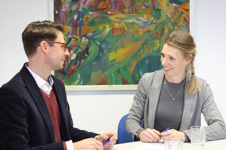 A woman and a man, both dressed in blazers, sit next to each other at a table and talk. A large colorful picture hangs on the wall in the background.