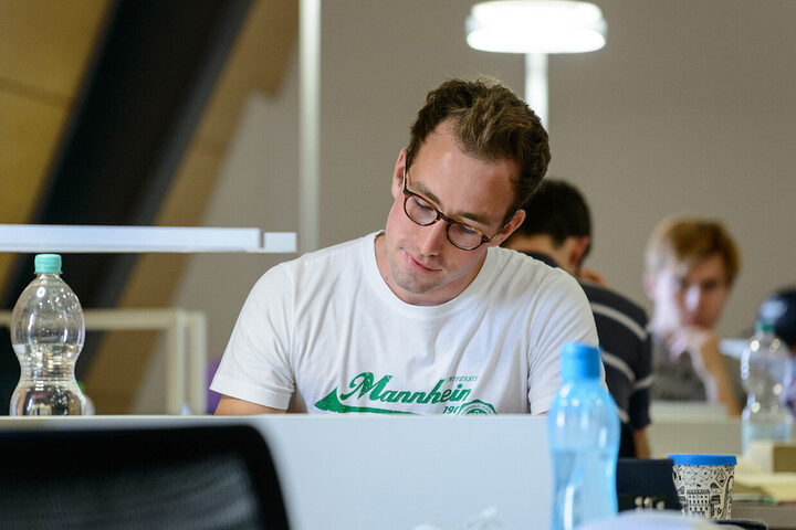 Ein Student mit Brille und einem weißen T-Shirt mit dem Schriftzug "Mannheim" sitzt an einem Arbeitsplatz in einer Bibliothek.