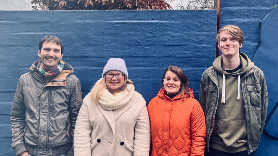 Außenaufnahme vor A5 von 4 RAM-Mitgliedern vor einer blauen Wand. 