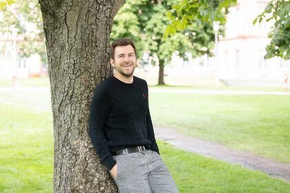 Tim Neubauer. Er lehnt locker an einem Baum und lacht, dabei trägt er einen schwarzen Pullover.
