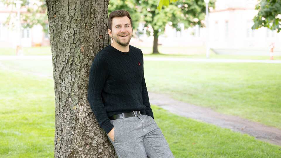 Tim Neubauer. Er lehnt locker an einem Baum und lacht, dabei trägt er einen schwarzen Pullover.