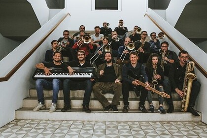 Eine Band (insgesamt 20 Personen) sitzt mit ihren Instrumenten auf der Treppe eines Universitätsgebäudes. Die Bandmitglieder halten ihre Instrumente so, als würden sie gerade darauf spielen.