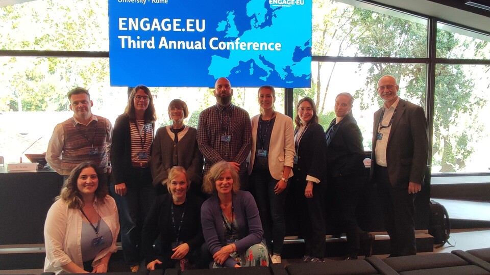 Eine Gruppe von Personen steht nebeneinander vor einem Fenster. Hinter ihnen hängt ein Banenr mit der Aufschrift "Engage.Eu - Third Annual Conference".