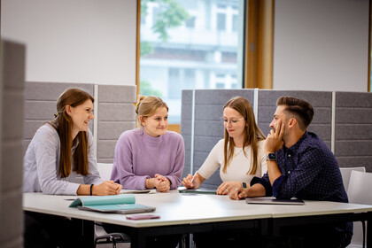 Vier junge Erwachsene sitzen in einem modernen Büro an einem Tisch und diskutieren konzentriert miteinander, vor ihnen liegen Notizbücher und Tablets.