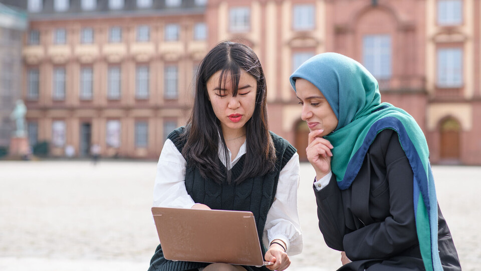 Zwei internationale Forscherinnen sitzen auf dem Ehrenhof vor dem Schloss und blicken auf einen Laptopbildschirm