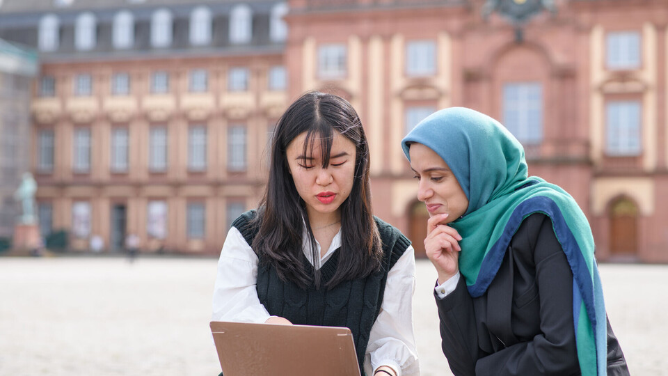 Zwei internationale Forscherinnen sitzen auf dem Ehrenhof vor dem Schloss und blicken auf einen Laptopbildschirm