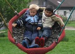 In einer großen Schaukel sitzen zwei Jungen Arm in Arm und halten sich fest, einer der Jungen ist blond und der andere ist schwarzhaarig.