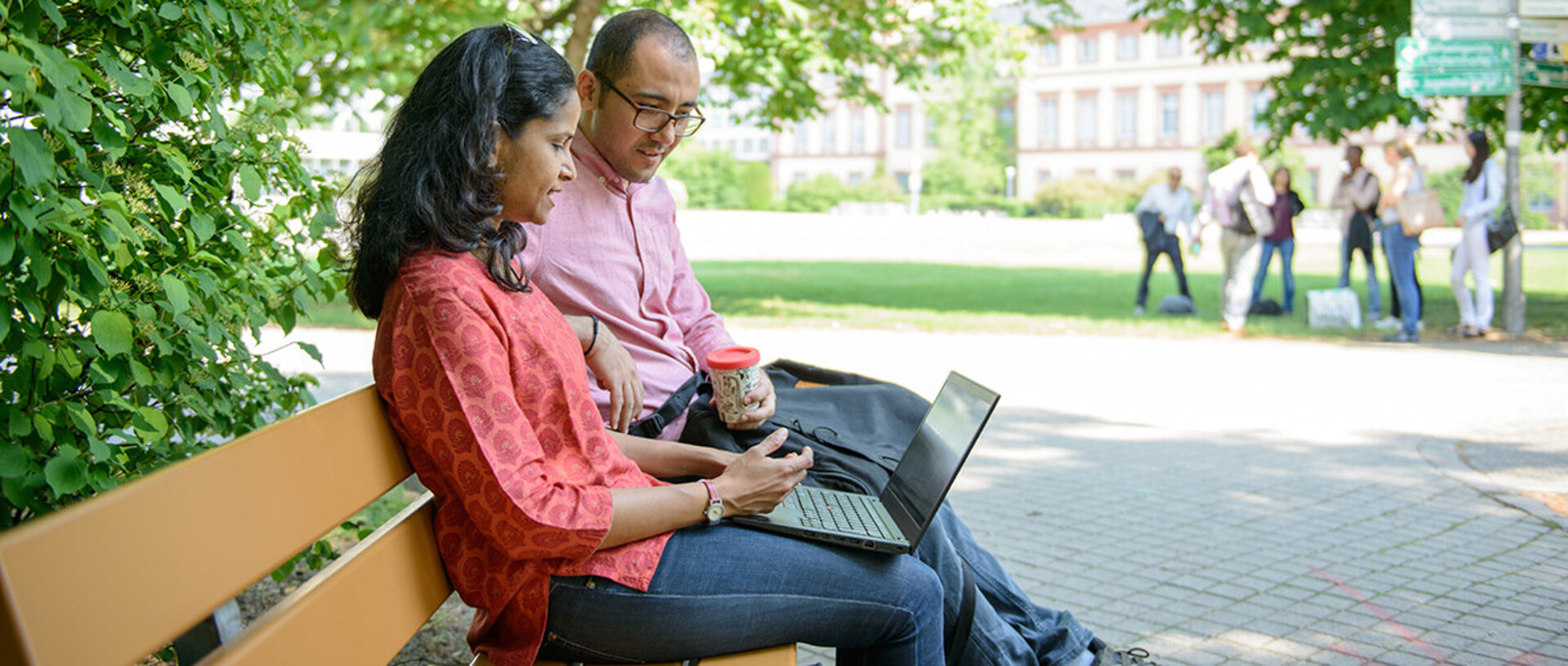 Zwei Personen sitzen mit einem Laptop auf einer Bank im Grünen.
