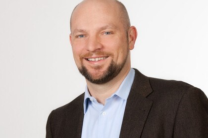 Eine lächelnde Person trägt ein hellblaues Hemd sowie einen braunen Blazer und steht vor einer weißen Wand. Die Person heißt Ulrich Wagner.