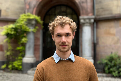 Max Otto. Er trägt einen braunen Pullover und steht vor einem alten Gebäude.