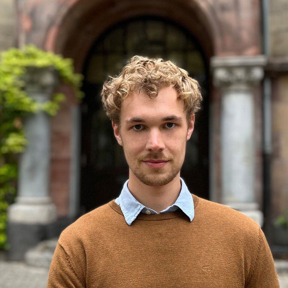 Max Otto. Er trägt einen braunen Pullover und steht vor einem alten Gebäude.