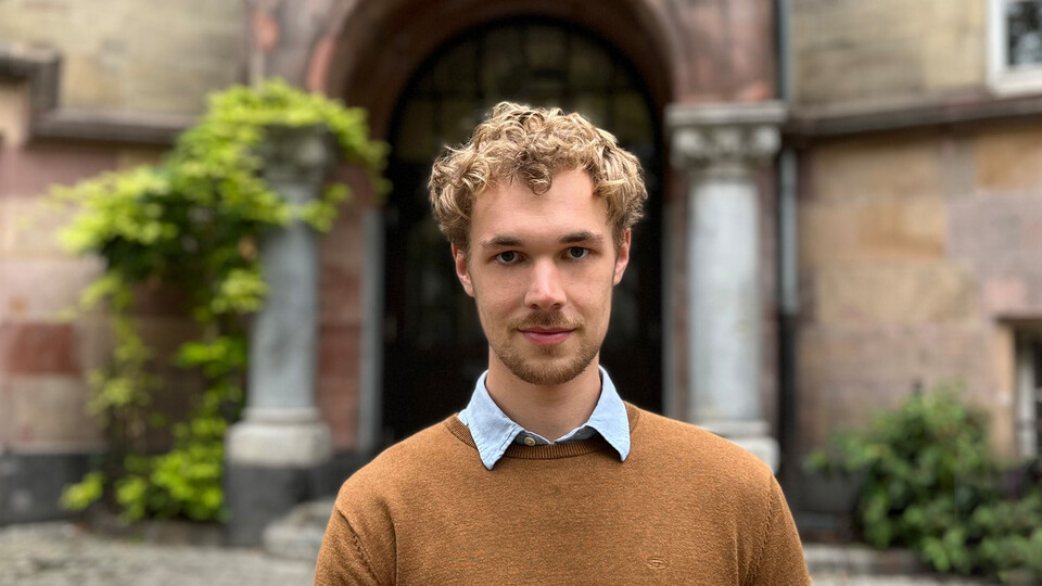 Max Otto. Er trägt einen braunen Pullover und steht vor einem alten Gebäude.