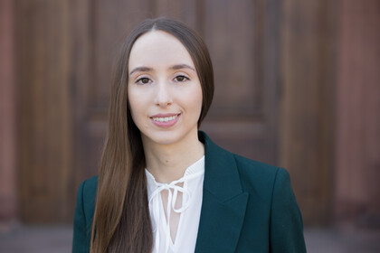 Sabine Stillger. Sie trägt ein graues Jackett über einem weißen Oberteil und steht vor einer Holztür.
