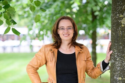 Portrait von Michele Tertilt. Sie trägt ein schwarzes Oberteil sowie eine orange-farbige Jacke und steht neben einem Baum auf einer Grünfläche.