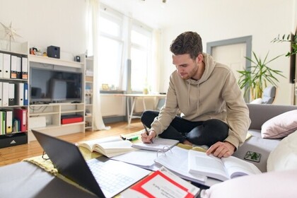 Studierender mit kurzen braunen Haaren und einem beigen Kapuzenpullover sitzt auf einem Sofa. Vor ihm liegen viele Bücher und ein Laptop. Er macht sich Notizen.