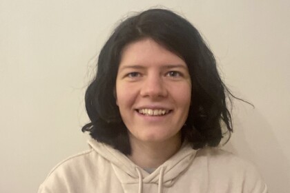 Lea Cohausz hat schulterlange schwarze Haare. Sie trägt einen cremefarbenen Kapuzenpullover und steht vor einer hellen Wand.