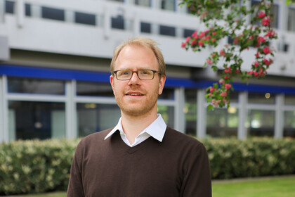 Portrait von Jochen Gebauer. Er trägt ein weißes Hemd sowie einen braunen Pullover und steht lächelnd vor dem A5-Gebäude.