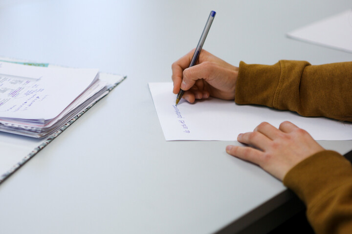 Eine Person schreibt mit einem Kugelschreiber auf ein Blatt Papier. Vor ihr liegt ein aufgeklappter Ordner.