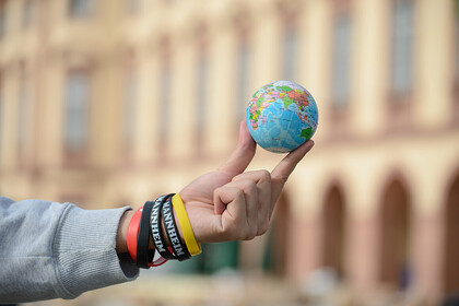 Hintergrund: Schloss Uni Mannheim, Vordergrund: ein Arm ragt ins Bild, zwiscen den Fingern wird ein Mini-Globus präsentiert