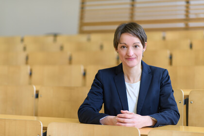 Portrait von Tabea Bucher-Koenen. Sie trägt eine weiße Bluse sowie einen dunkelblauen Blazer und sitzt mit gefaltetenen Händen in einem leeren Hörsaal.