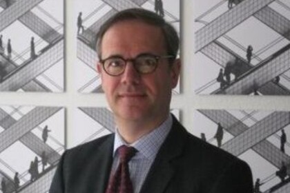 Portrait von Bernhard Ebbinghaus. Er trägt ein wißes Hemd sowie eine rote Krawatte und einen schwarzen Blazer. Er steht lächelnd vor einer Bilderwand.