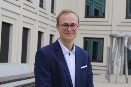 Thilo Dieing steht vor einem Gebäude der Universität. Er hat blonde Haare und trägt eine Brille, ein weißes Hemd und einen dunkelblauen Anzug.