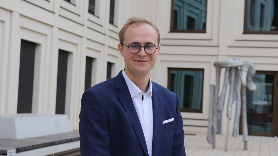 Thilo Dieing steht vor einem Gebäude der Universität. Er hat blonde Haare und trägt eine Brille, ein weißes Hemd und einen dunkelblauen Anzug.