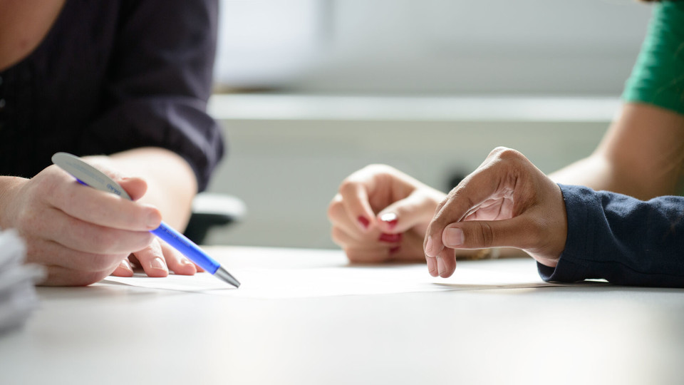 Internationale Studierende Zwei Studierende werden beraten, dabei sind die Hände und der Stift der Mitarbeiterin, der auf ein Papier zeigt, im Fokus.