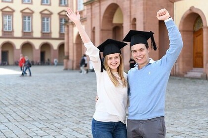 Zwei Studierende tragen einen Absolventenhut auf dem Kopf und strecken einen Arm in die Luft. Im HIntergrund ist die Universität Mannheim zu sehen