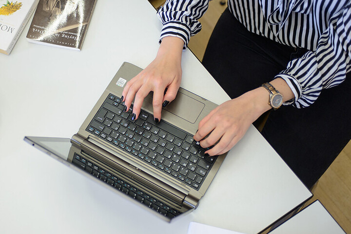 Eine Person tippt auf einem Laptop. Ihre Hände sind auf der Tastatur zu sehen, und sie trägt eine gestreifte Bluse.